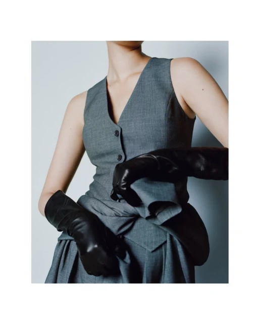 Fashion photo of a woman wearing a grey vest and skirt with black leather gloves against a light grey background in a studio setting.