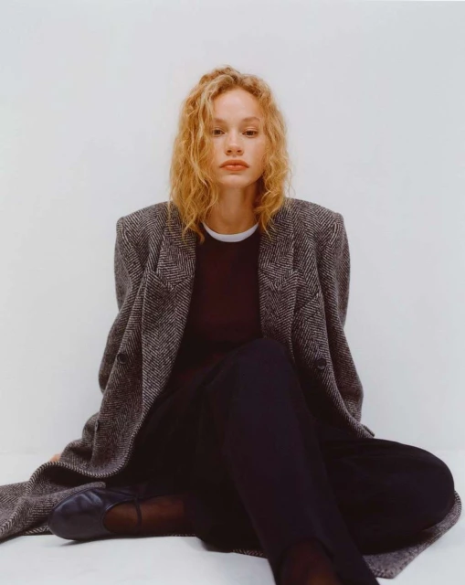 A blonde woman with curly hair sits in a studio wearing a herringbone coat, black top, and pants, looking at the camera.