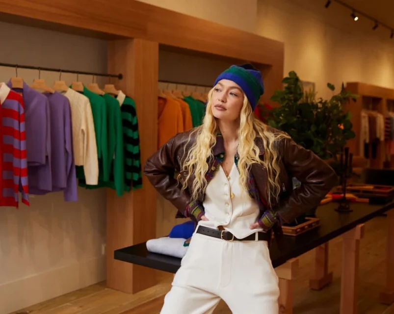 A blonde woman models a beanie, leather jacket, and white pantsuit in a clothing store with racks of colorful sweaters and tops.