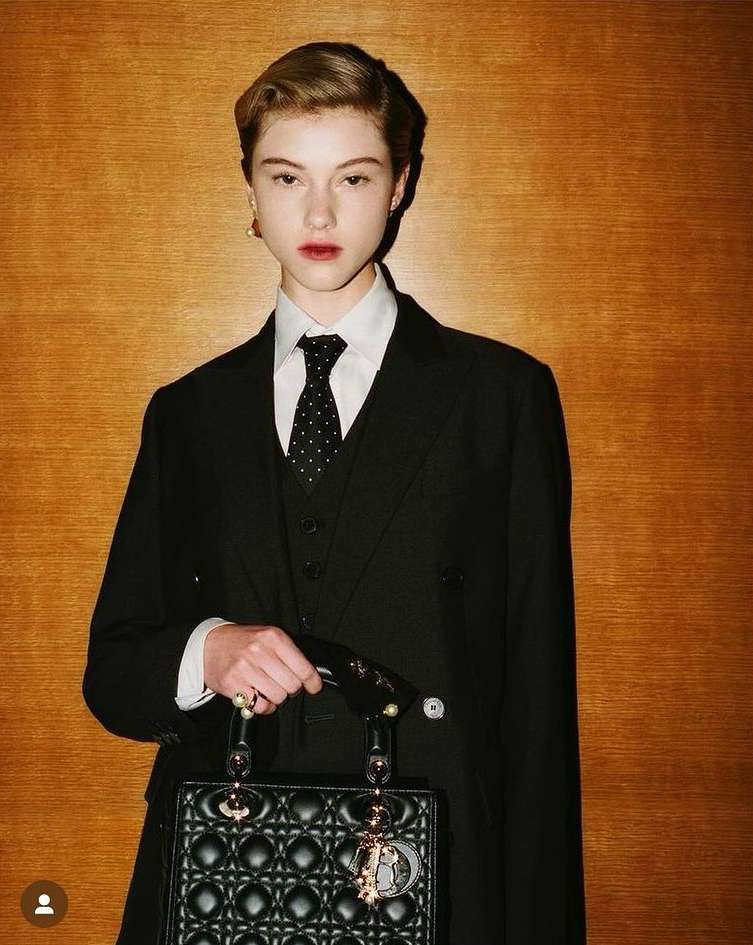 Elegant young woman in a black suit and tie holds a black quilted purse against a warm wood background, in a fashion-forward editorial style.