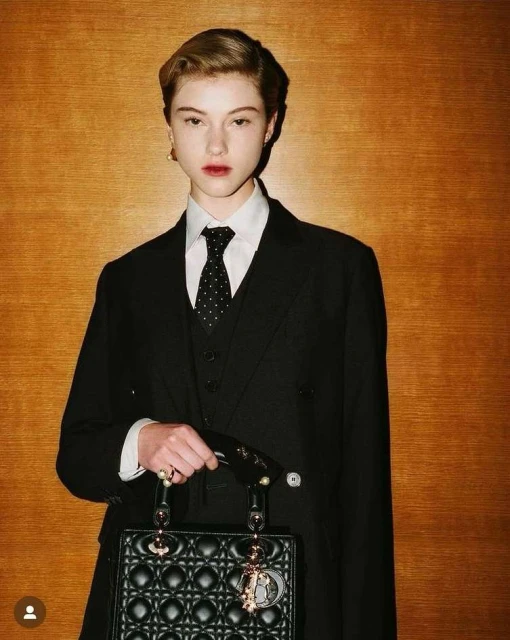 Elegant young woman in a black suit and tie holds a black quilted purse against a warm wood background, in a fashion-forward editorial style.