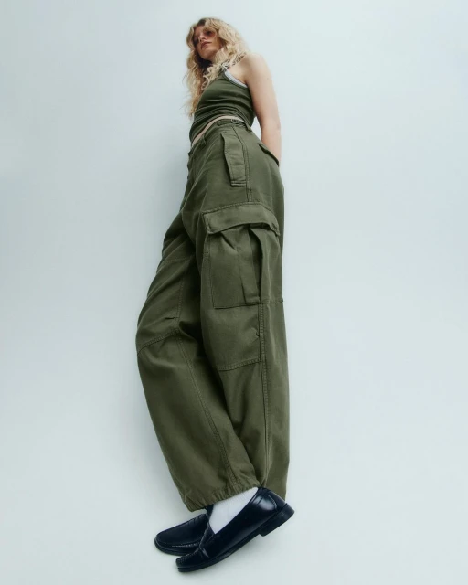A stylish adult female model is shot from a low angle wearing a green crop top and wide-leg cargo pants against a white background.