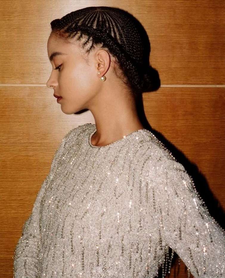 Profile portrait of a young adult woman with braided hair wearing a beaded top against a wood panel background.