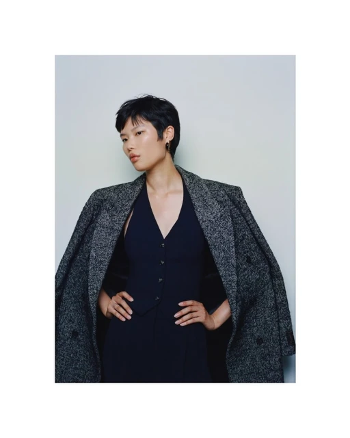 Fashion portrait of an Asian woman in a navy dress and gray wool coat, gazing to the side with a neutral expression on a light background.