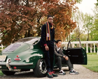 Two men posing next to a vintage dark green sports car parked on a gravel path with trees and classical colonnade in the background.
