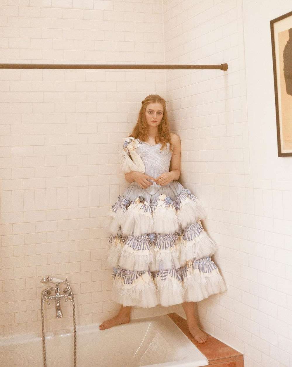 A red-haired woman posing barefoot in a white and blue frilled gown inside a white tiled bathroom with antique-style fixtures and a framed print.