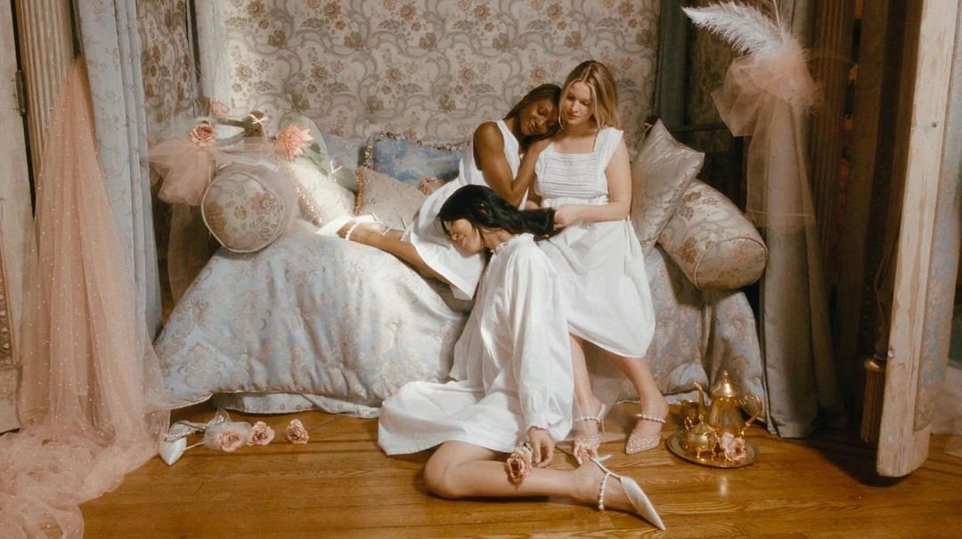 Three women in white dresses pose together indoors with soft, romantic lighting and floral accents for a fashion or beauty editorial.