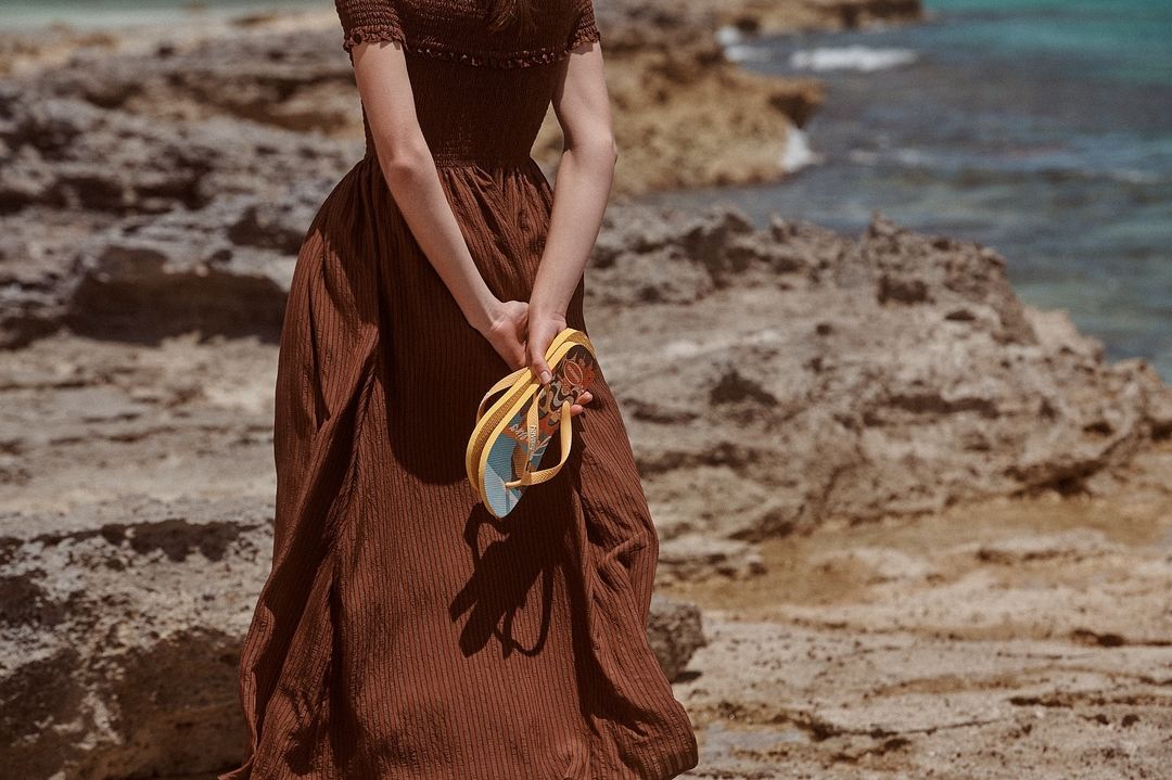 A woman in a brown dress stands on a rocky beach near turquoise ocean water, holding yellow flip-flops behind her back.