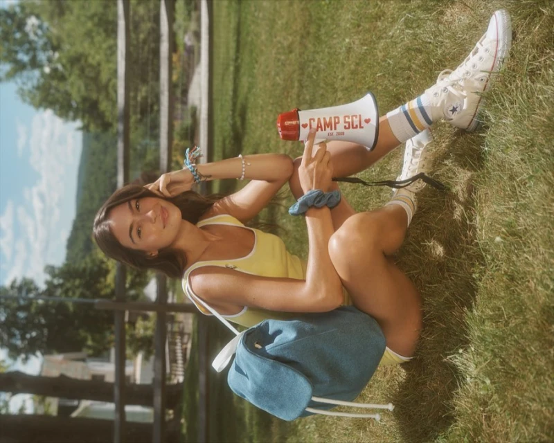 Young woman sits on grass with a megaphone, wearing a yellow top, sneakers, and socks in a sun-drenched outdoor environment.