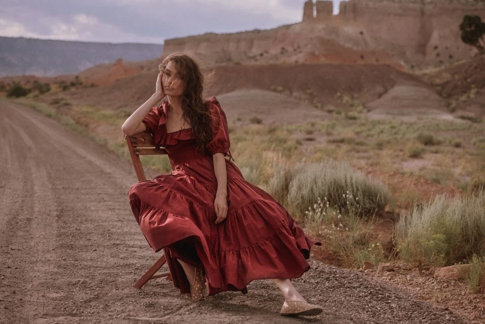 Adult woman in red dress sits on chair on dirt road with desert landscape and rock formations in the background, under cloudy sky.