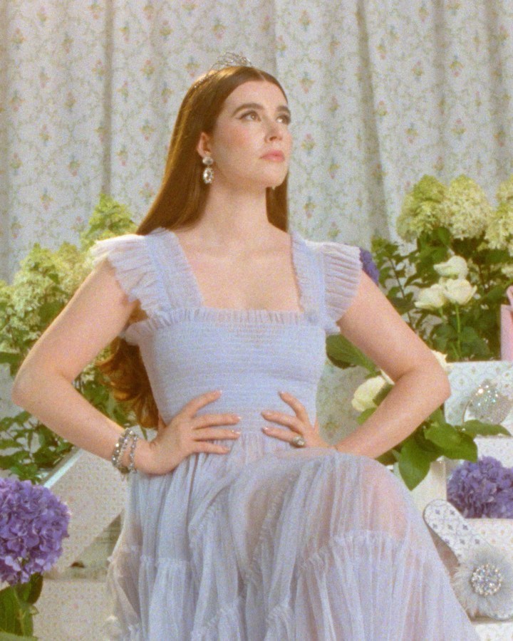 Woman wearing a tiara and light purple dress sitting with her hands in her lap with flowers and floral wallpaper behind her.