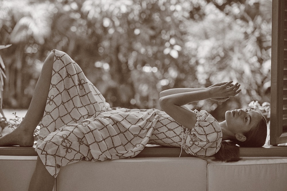 A woman in a patterned dress is lying down on a window seat, with trees in the background in this sepia-toned outdoor fashion shot.