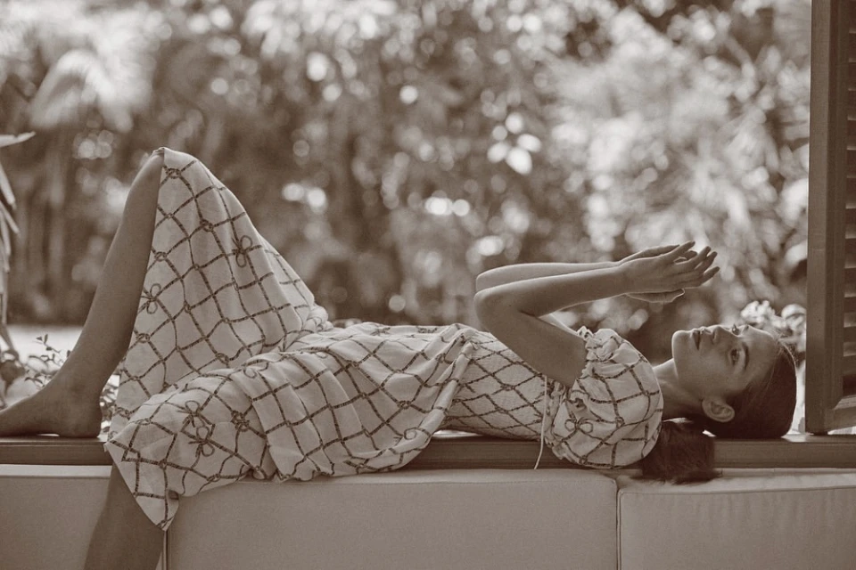 A woman in a patterned dress is lying down on a window seat, with trees in the background in this sepia-toned outdoor fashion shot.