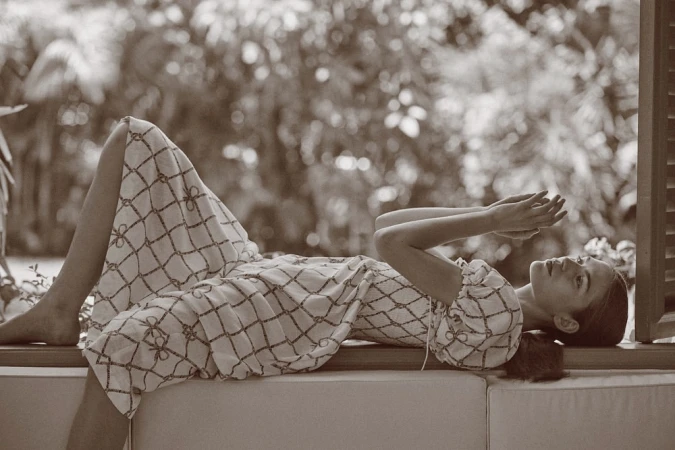 A woman in a patterned dress is lying down on a window seat, with trees in the background in this sepia-toned outdoor fashion shot.