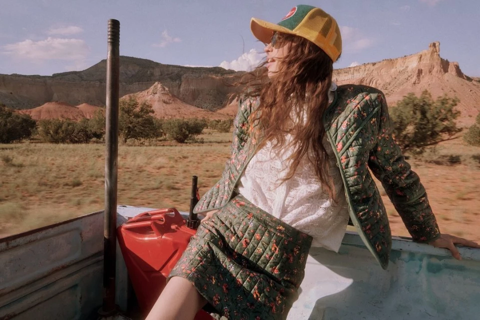 A woman in a floral jacket and skirt sits in the back of a truck with mountains in the background on a sunny day outdoors.