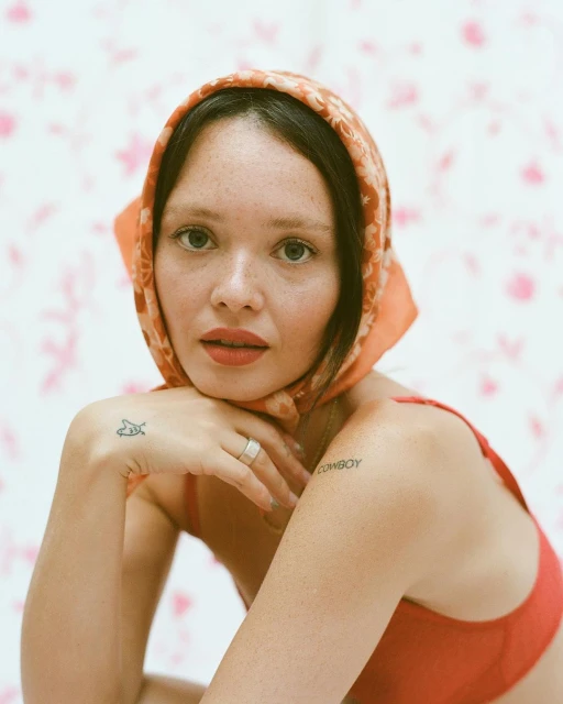 Woman wearing an orange headscarf and red bra top poses indoors against a pink floral background in a medium, eye-level shot.
