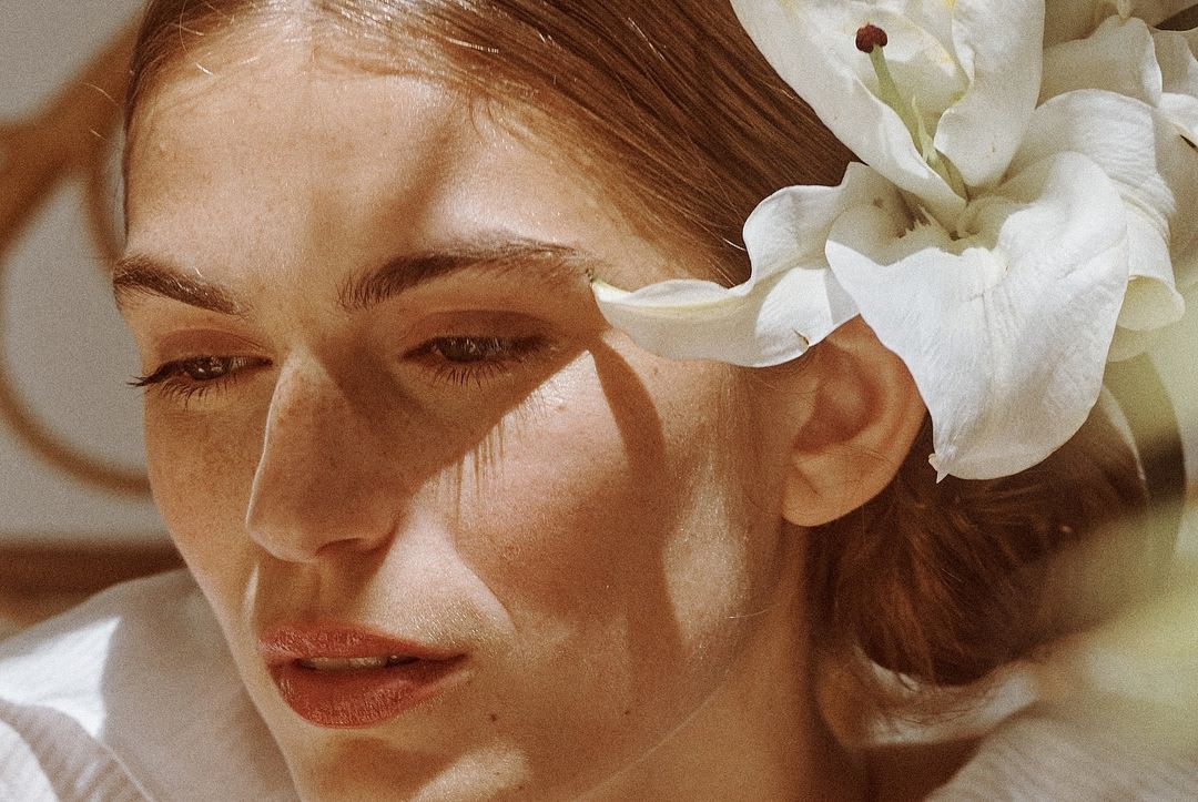 Close-up portrait of a fair-skinned woman with light brown hair styled with white lily, looking down in an indoor environment with soft lighting.