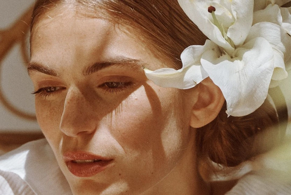 Close-up portrait of a fair-skinned woman with light brown hair styled with white lily, looking down in an indoor environment with soft lighting.