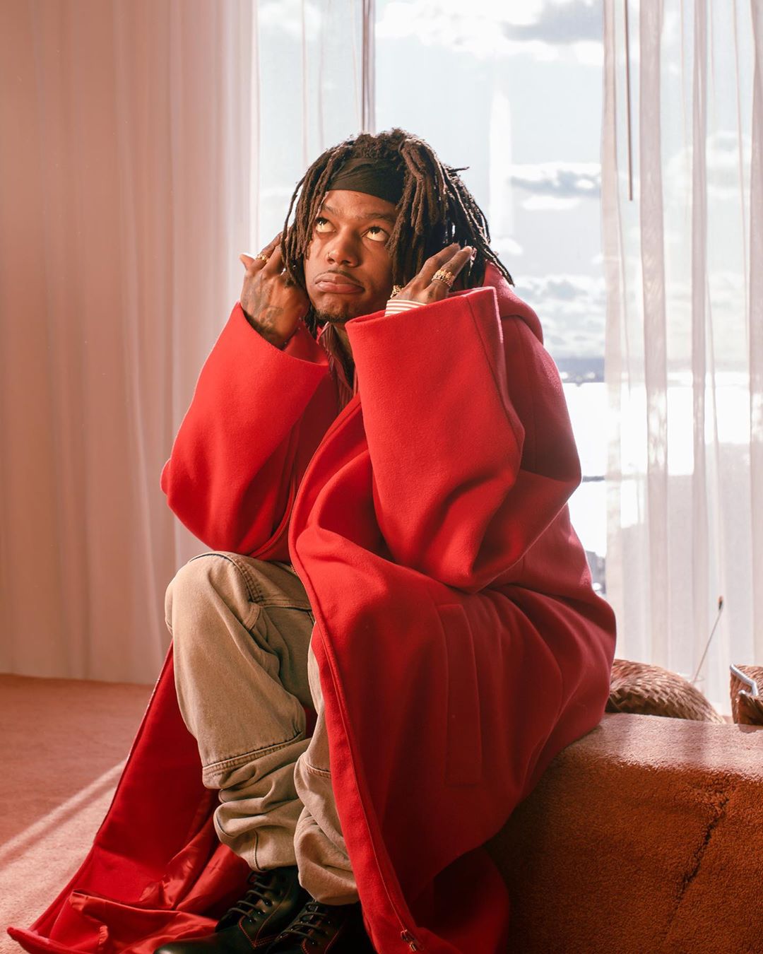 Adult male with dreadlocks wearing a red robe and black shoes sits on a sofa indoors near a bright window with white curtains.