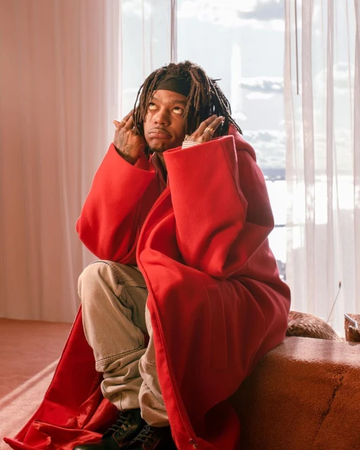 Adult male with dreadlocks wearing a red robe and black shoes sits on a sofa indoors near a bright window with white curtains.