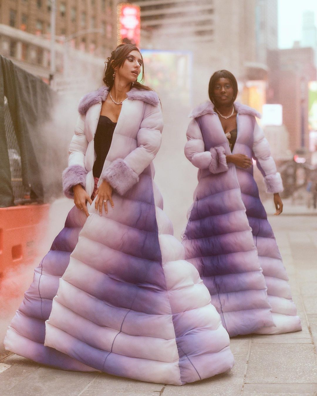 Two women wearing long ombre puffer coats standing on a city street with atmospheric haze.
