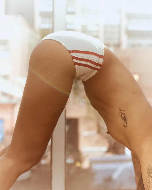 Torso shot of a woman in white bikini bottoms with red stripes, back arched with tattoos visible, against a blurred city background.