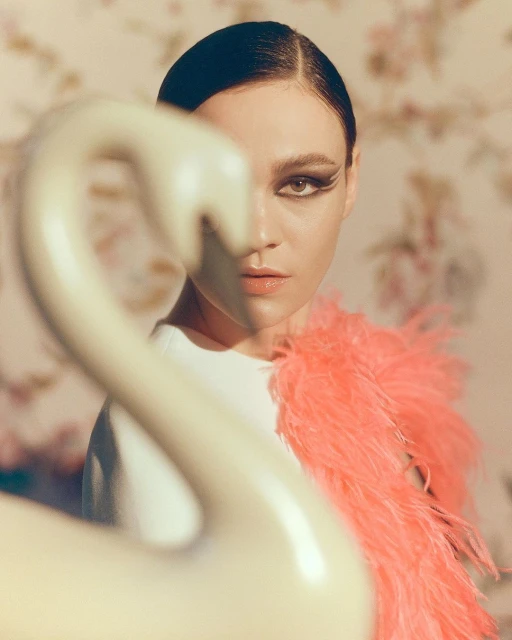 Portrait of a woman with dramatic eye makeup and a coral feather boa, standing in front of a vintage floral wallpaper.