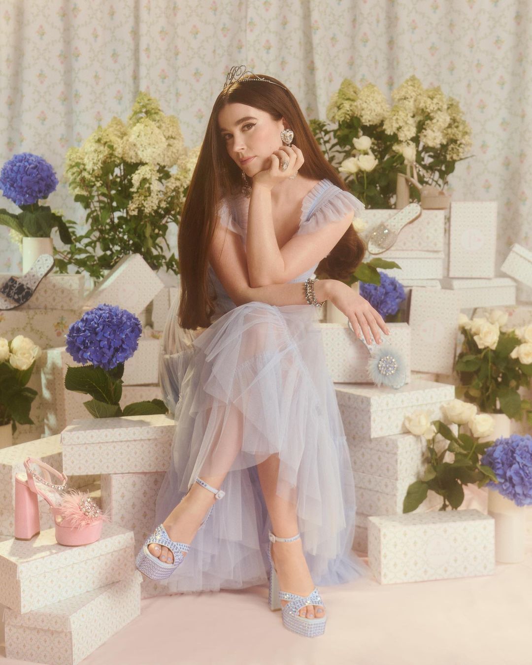 A woman wearing a crown and a light blue dress and platform heels sits among gift boxes and white and blue hydrangeas.