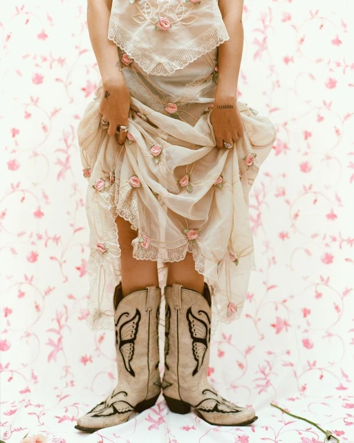 A person in a dress with floral details and cowboy boots stands against a pink floral background, creating a vintage fashion aesthetic.