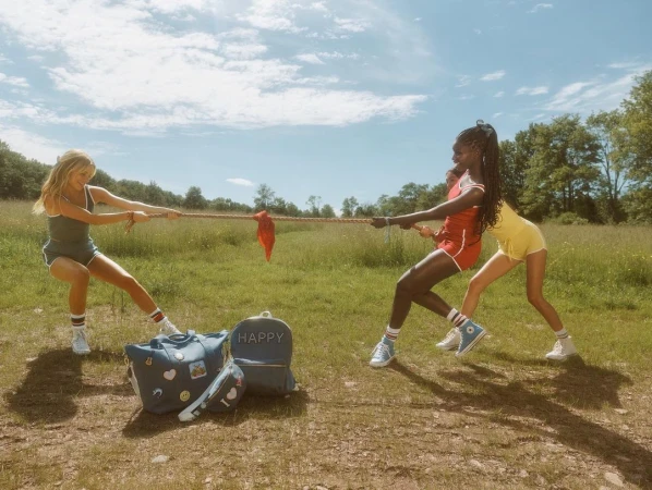 Three women engage in a tug-of-war on a grassy field under a blue sky, with backpacks and bags in front, capturing a playful outdoor scene.