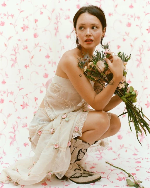 A woman in a light-colored dress squats and holds a bouquet against a floral background, wearing boots with embroidered designs.