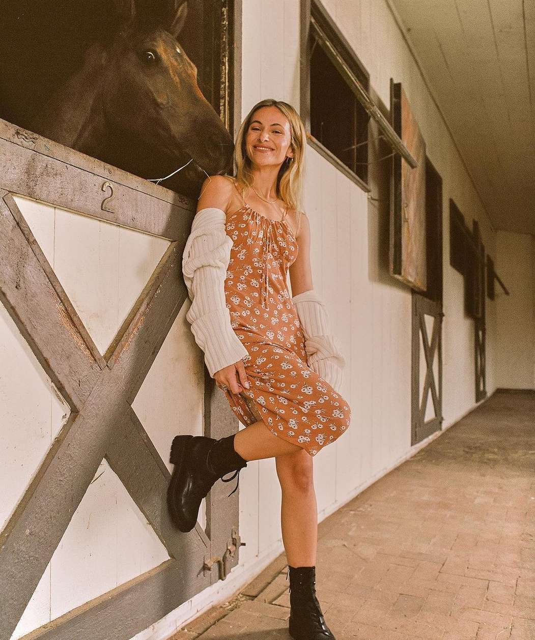 Woman in floral dress leans against stable with a horse peering from a stall, wearing boots and cardigan in a candid setting.