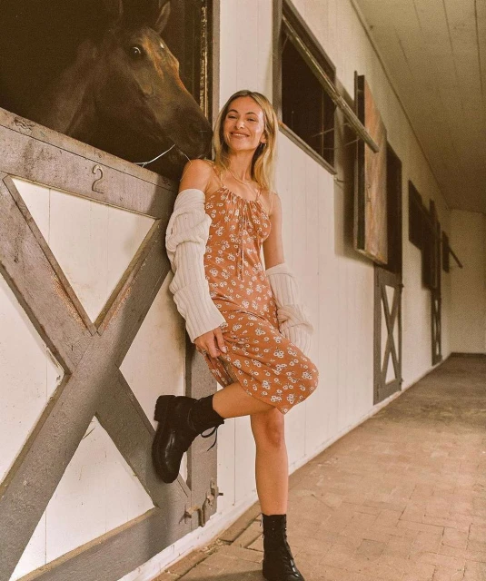 Woman in floral dress leans against stable with a horse peering from a stall, wearing boots and cardigan in a candid setting.
