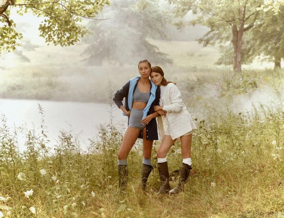 Two female models posing by a calm lake in a misty meadow wearing knitwear and tall boots, photographed by Emma Craft.