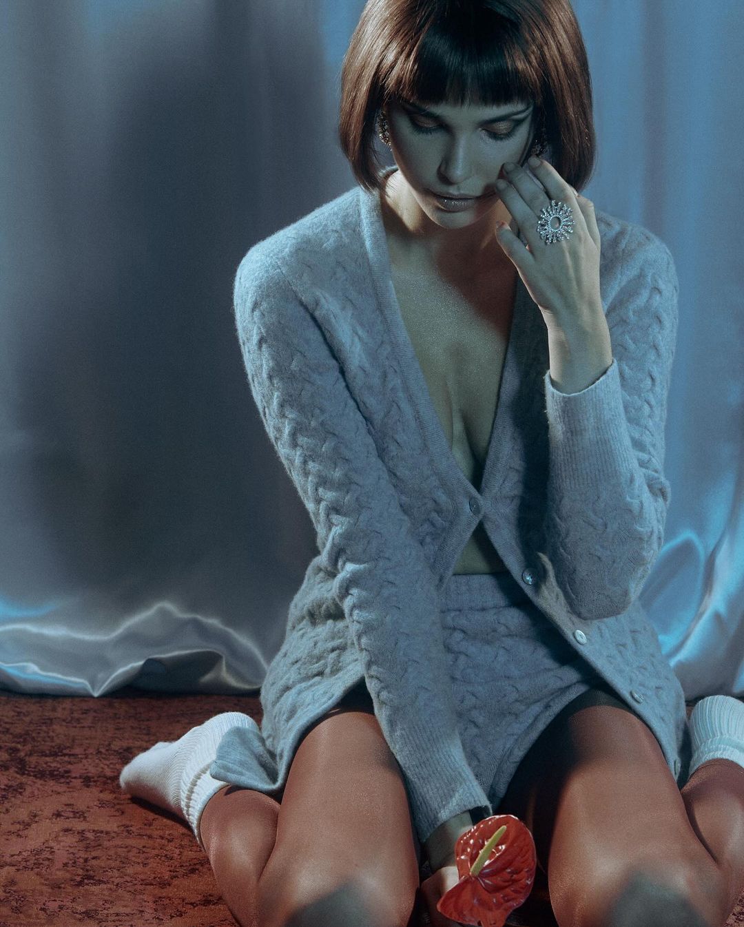 Brunette woman poses in a matching knit cardigan and shorts set while kneeling on an orange rug, against a hazy gray backdrop.