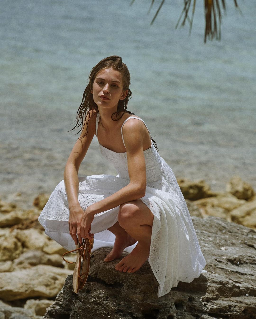 Young woman in white dress poses on a rock near a tropical beach with blue water, holding sandals and wearing a summer dress.