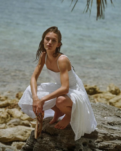 Young woman in white dress poses on a rock near a tropical beach with blue water, holding sandals and wearing a summer dress.