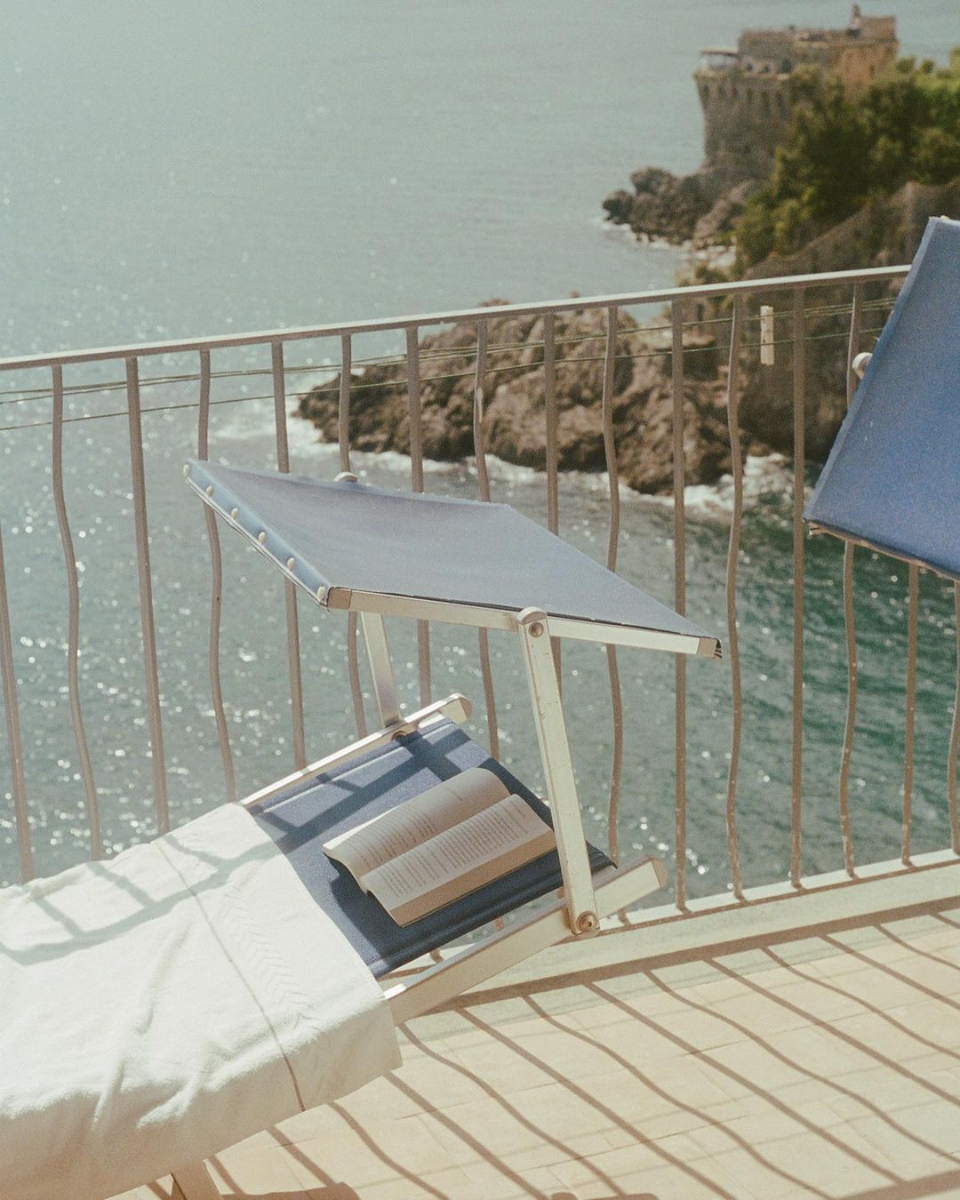 Coastal scene with sun loungers on a balcony overlooking the ocean, featuring a stone building on a rocky outcrop.