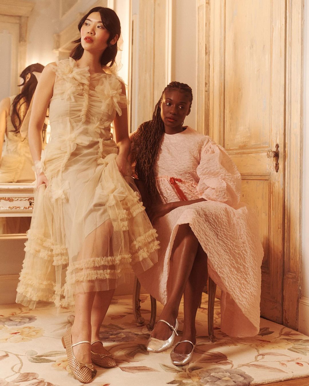 Two female models in textured dresses pose in an interior room with wooden doors and a floral rug, photographed by Emma Craft.