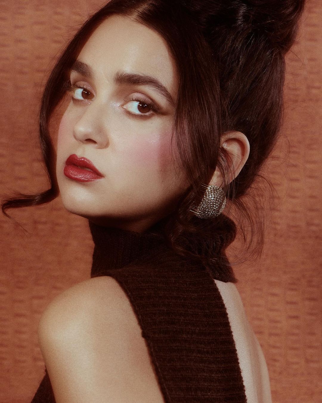 Headshot of a woman with dark hair in a high bun, wearing a brown turtleneck dress, and statement earrings against a textured background.