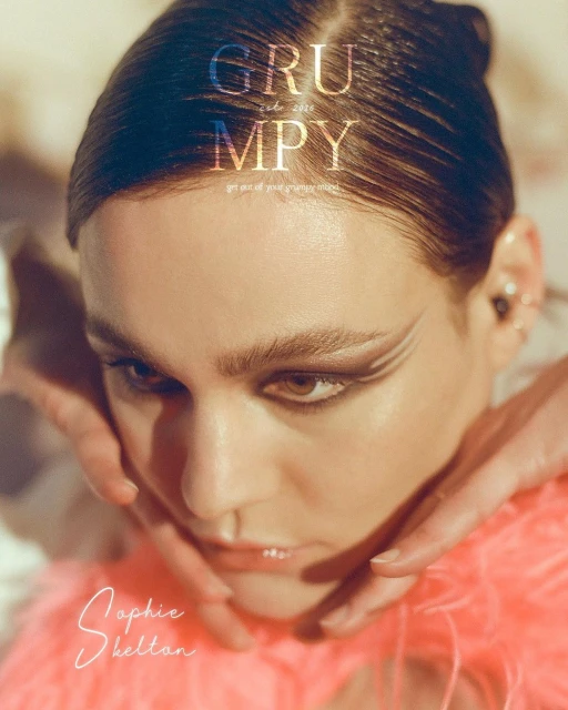 Close-up portrait of a woman with dramatic makeup and hands framing her face, set against a soft, neutral backdrop with light gradients.