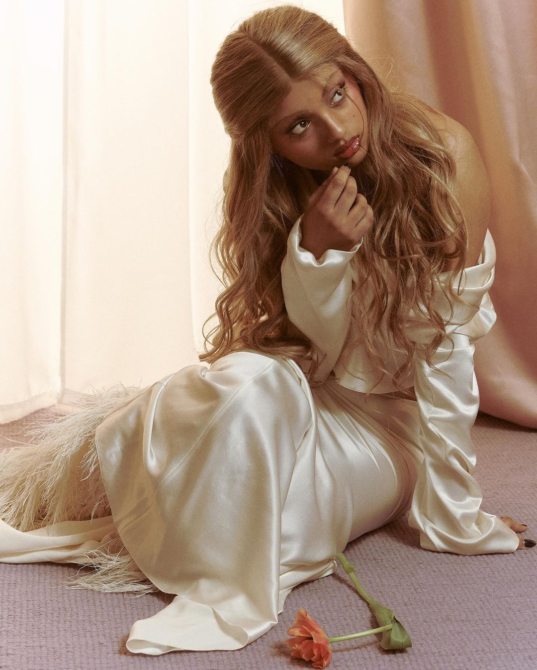 Portrait of a woman with wavy blonde hair and a satin dress, seated indoors with a flower lying on the carpet, gazing upwards.