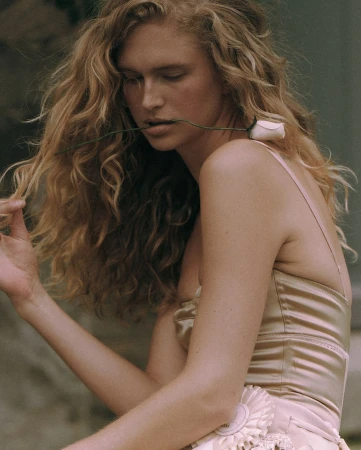 Woman with long blonde curly hair holding a rose in her mouth wearing a satin top