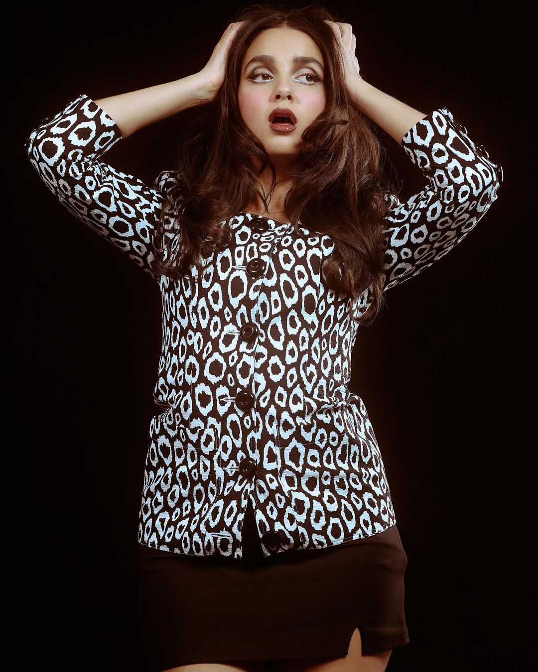 Woman with surprised expression posing in leopard-print jacket and black skirt against dark backdrop with hands on her head.