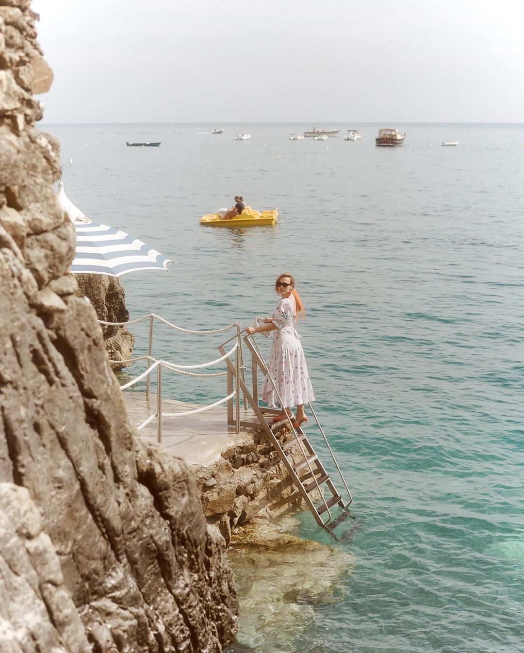 Woman in floral dress descends metal stairs into ocean water. Cliffside location with boats visible in background on sunny day.