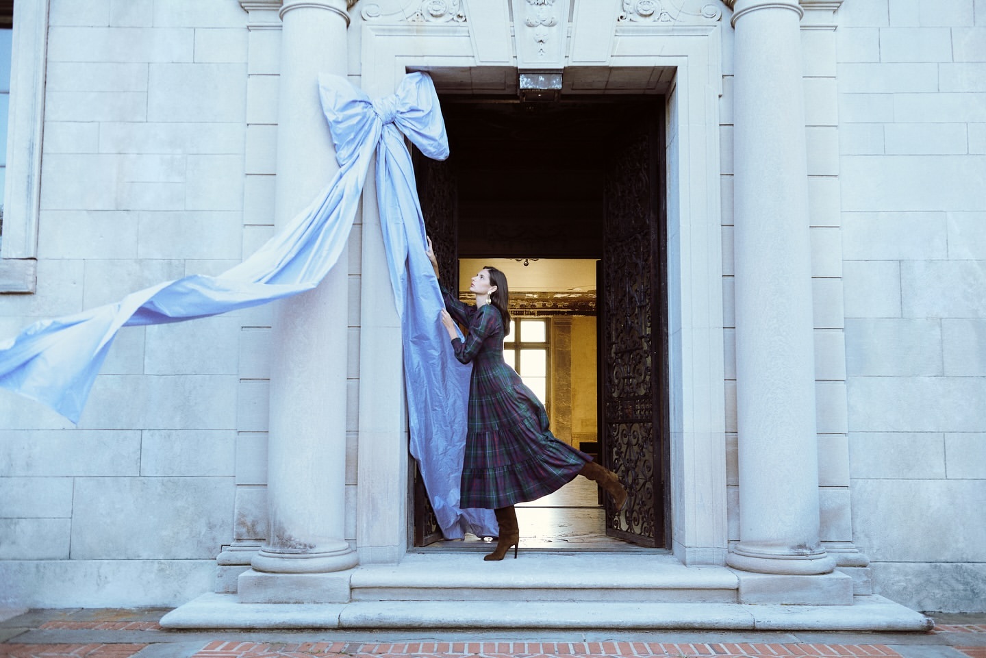 A woman poses in a plaid dress with a large blue bow attached to the stone entrance of a building, creating a striking visual contrast.