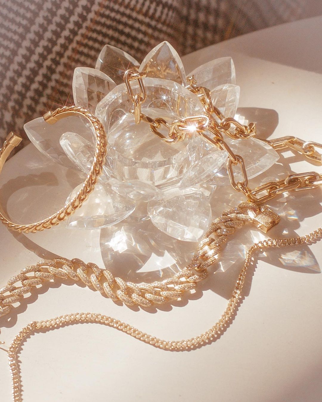 Close-up of gold jewelry, including chains and a cuff bracelet, arranged on a transparent decorative object, under soft, natural light.