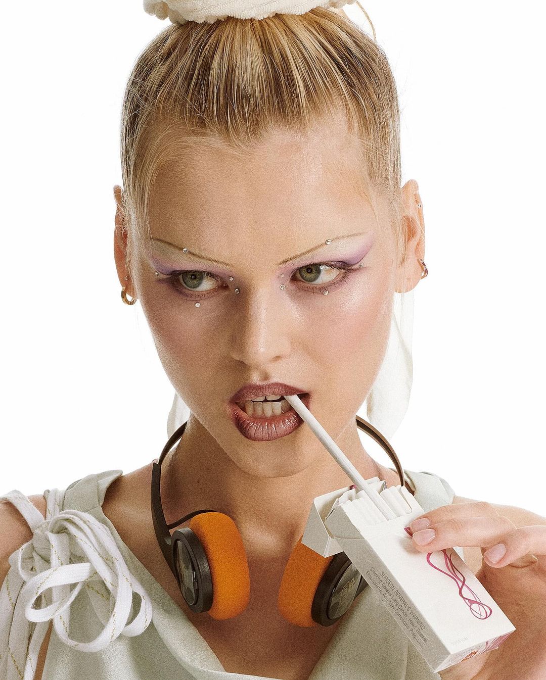 Close-up of a woman with painted eyebrows and headphones, holding a cigarette in her mouth and a pack of cigarettes in her hand.
