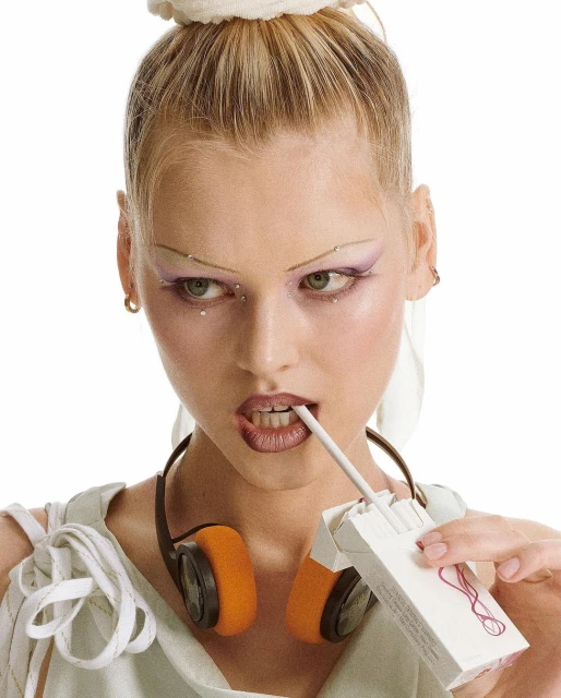 Close-up of a woman with painted eyebrows and headphones, holding a cigarette in her mouth and a pack of cigarettes in her hand.