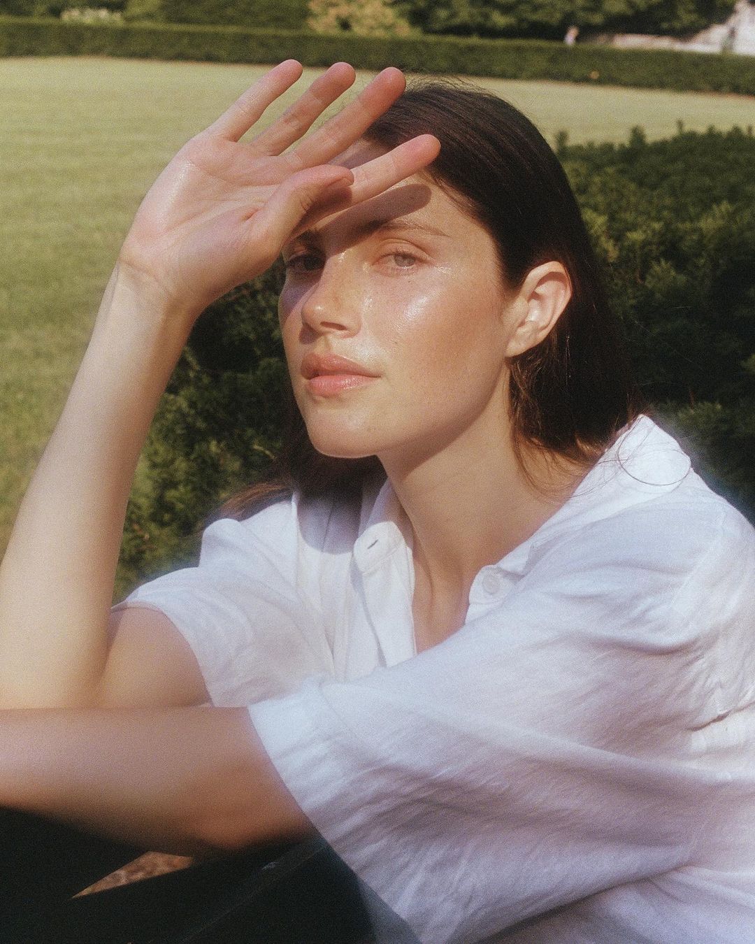Portrait of a young woman with brown hair shielding her eyes in an outdoor setting, wearing a white collared shirt.